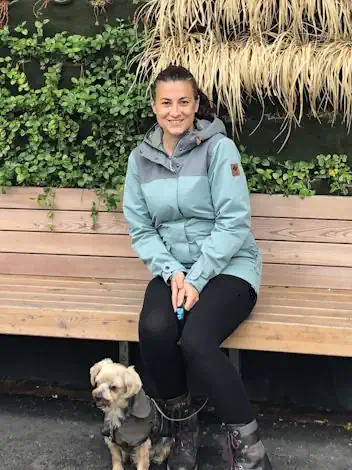 Laura Bisaccia, B.Sc., Psychologische Beraterin in Graz, Steiermark, Österreich und Raaba, Graz Umgebung, Steiermark, Österreich sitzt auf einer hellen Holzbank im Freien. Im Hintergrund sind grüne Pflanzen und Gräser. Zu Ihre Füßen sitzt ein kleiner Hund.